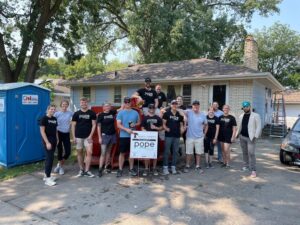 A group of volunteers wearing matching black "Pope Design Group" t-shirts pose in front of a single-story house with white brick exterior and a brown shingled roof. The team is smiling and standing in front of a red dumpster, with some members leaning on it. A sign in the foreground reads "Hearts & Hammers" and features the "Pope Design Group" logo. A portable toilet from "OnSite" is visible on the left, and a ladder is propped against the house on the right. The background includes green trees and a clear sky, indicating a sunny day. The group appears to be engaged in a home improvement or rehabilitation project.