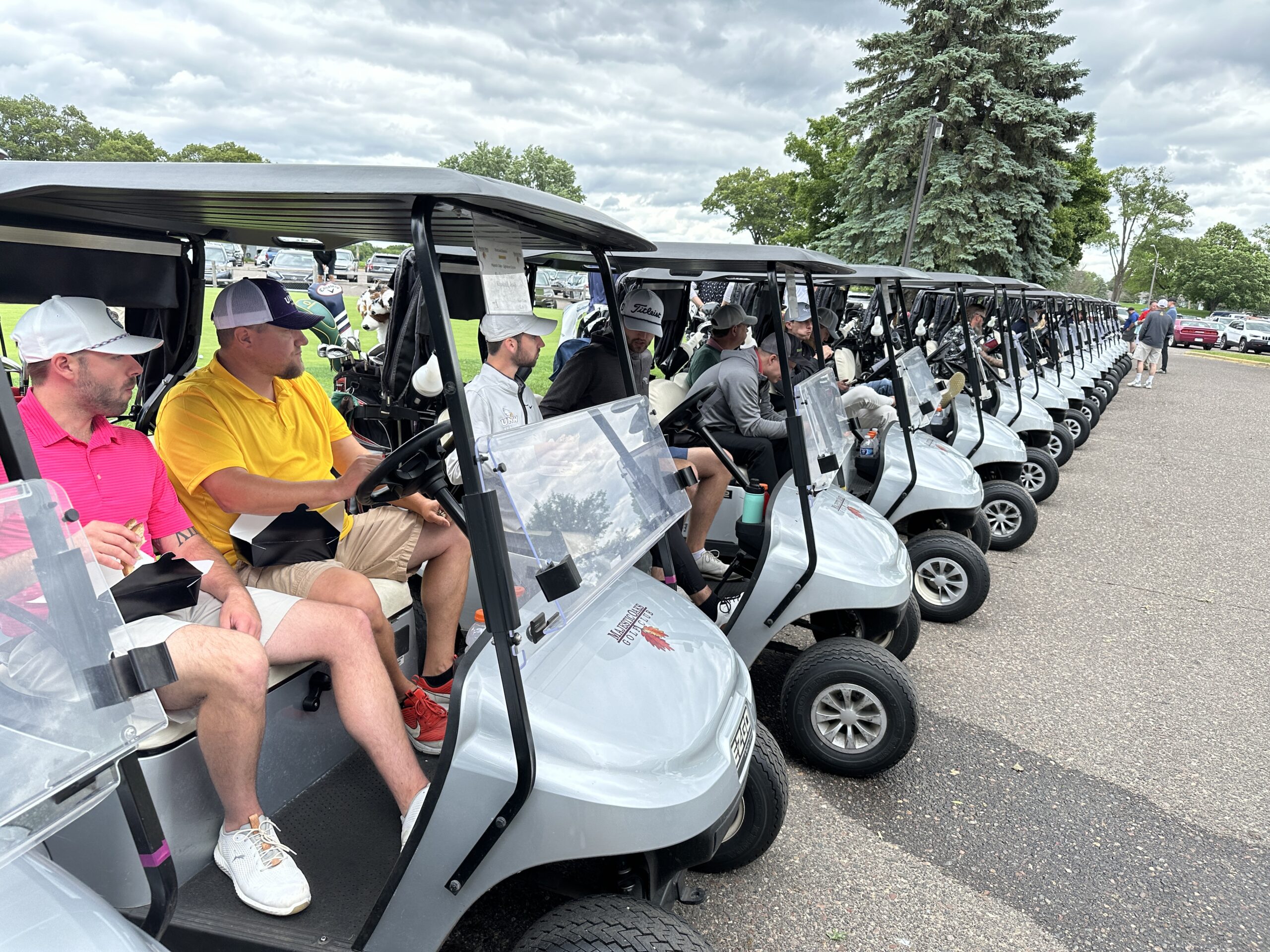 A row of golf carts lined up in a parking lot, each occupied by golfers preparing for an event. The participants, dressed in golf attire, are engaged in conversation and eating snacks while waiting. The sky is overcast, and trees, parked cars, and additional golfers walking in the distance are visible in the background.