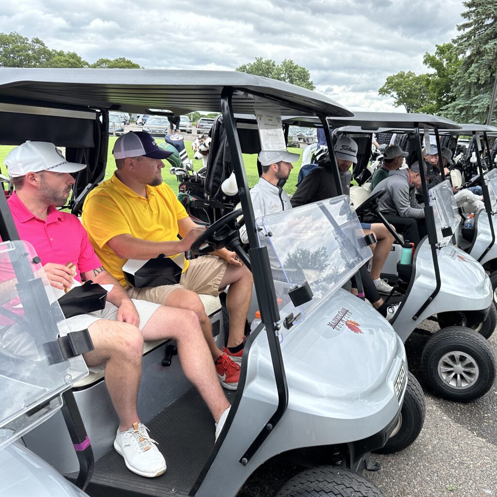 A row of golf carts lined up in a parking lot, each occupied by golfers preparing for an event. The participants, dressed in golf attire, are engaged in conversation and eating snacks while waiting. The sky is overcast, and trees, parked cars, and additional golfers walking in the distance are visible in the background.