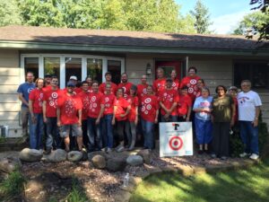 "A group of Hearts & Hammers volunteers in red Target-branded shirts posing in front of a single-story home they worked on. The team stands alongside the homeowners, smiling. A Hearts & Hammers sign with the Target logo is placed in the front yard, indicating sponsorship. The background features trees and a well-maintained lawn