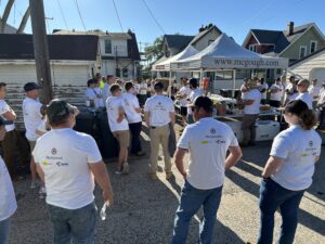 Alt text: A group of volunteers wearing McGough-branded white t-shirts gathers outdoors in a residential neighborhood for a community service event. They are listening to a speaker near a McGough-branded canopy tent, which has a table with refreshments. The setting includes houses, garages, and a utility pole, indicating a home improvement or renovation project. The weather is sunny and bright.