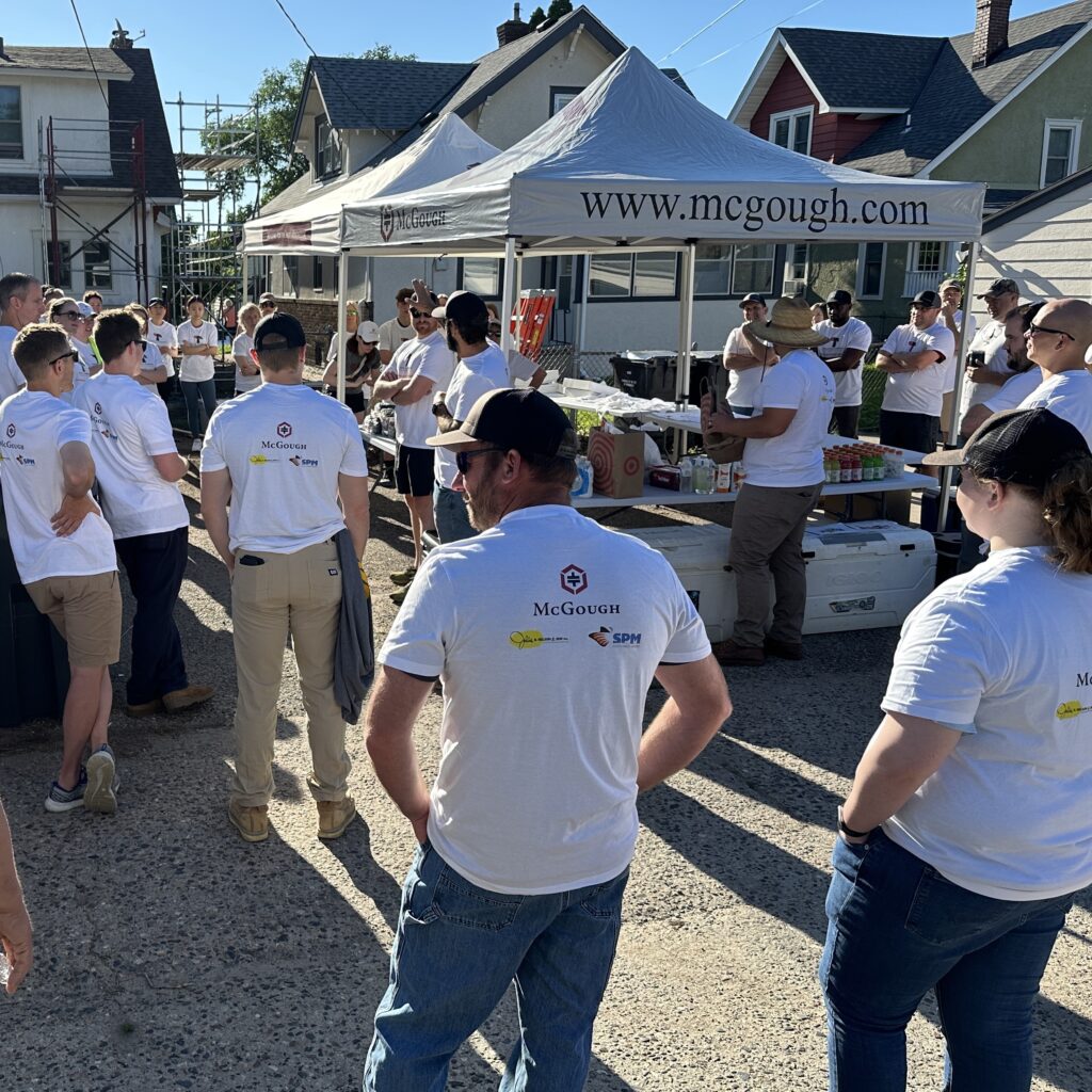 Alt text: A group of volunteers wearing McGough-branded white t-shirts gathers outdoors in a residential neighborhood for a community service event. They are listening to a speaker near a McGough-branded canopy tent, which has a table with refreshments. The setting includes houses, garages, and a utility pole, indicating a home improvement or renovation project. The weather is sunny and bright.