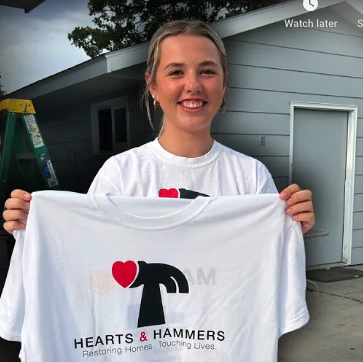 "Young volunteer smiling while holding up a Hearts & Hammers t-shirt in front of a recently restored home, showcasing the organization's mission to restore homes and touch lives."